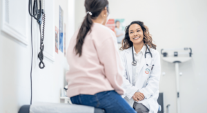 a middle aged woman sits up on an exam table as she talks with her female doctor about health concerns