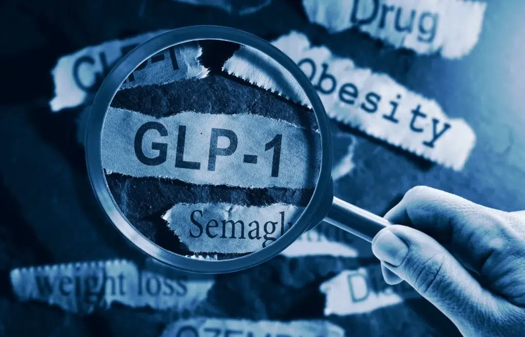 A close-up image shows a hand holding a magnifying glass over torn paper strips. The magnified text reads “GLP-1” and “Semaglutide.” Surrounding torn paper pieces display words such as “Obesity,” “weight loss,” “Drug,” and “Diabetes.” The overall image has a blue tint, symbolizing medical or pharmaceutical context.