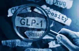 A close-up image shows a hand holding a magnifying glass over torn paper strips. The magnified text reads “GLP-1” and “Semaglutide.” Surrounding torn paper pieces display words such as “Obesity,” “weight loss,” “Drug,” and “Diabetes.” The overall image has a blue tint, symbolizing medical or pharmaceutical context.