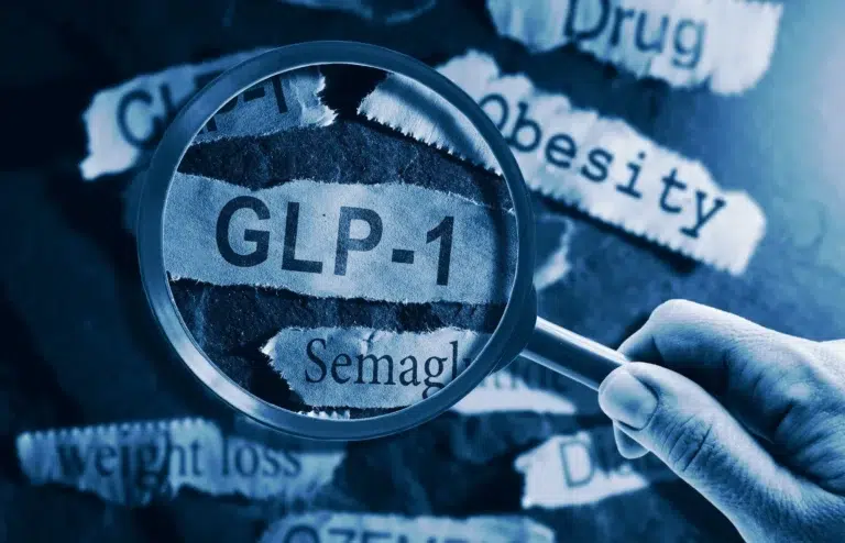 A close-up image shows a hand holding a magnifying glass over torn paper strips. The magnified text reads “GLP-1” and “Semaglutide.” Surrounding torn paper pieces display words such as “Obesity,” “weight loss,” “Drug,” and “Diabetes.” The overall image has a blue tint, symbolizing medical or pharmaceutical context.
