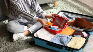 A woman sits on the floor beside an open suitcase filled with clothes, preparing for her travel health essentials.