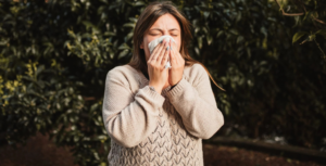 A woman blowing her nose in front of a tree, illustrating symptoms related to nasal drip cough and its medical implications.