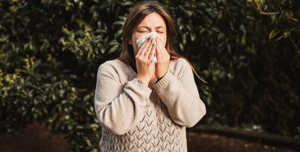A woman blowing her nose in front of a tree, illustrating symptoms related to nasal drip cough and its medical implications.