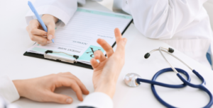 A doctor records notes on a clipboard as a patient watches, indicating a consultation in a medical office.